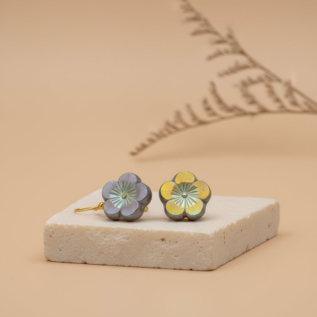 A pair of flower-shaped dangle earrings with gold-tone hooks and petals, featuring iridescent gray-blue outer petals and yellow inner centers, displayed against a light gray background.