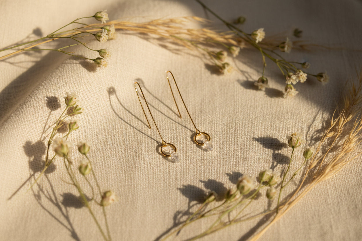 A pair of gold-toned dangle earrings with thin vertical bars ending in small circular hoops, displayed on a clear stand against a light gray background.