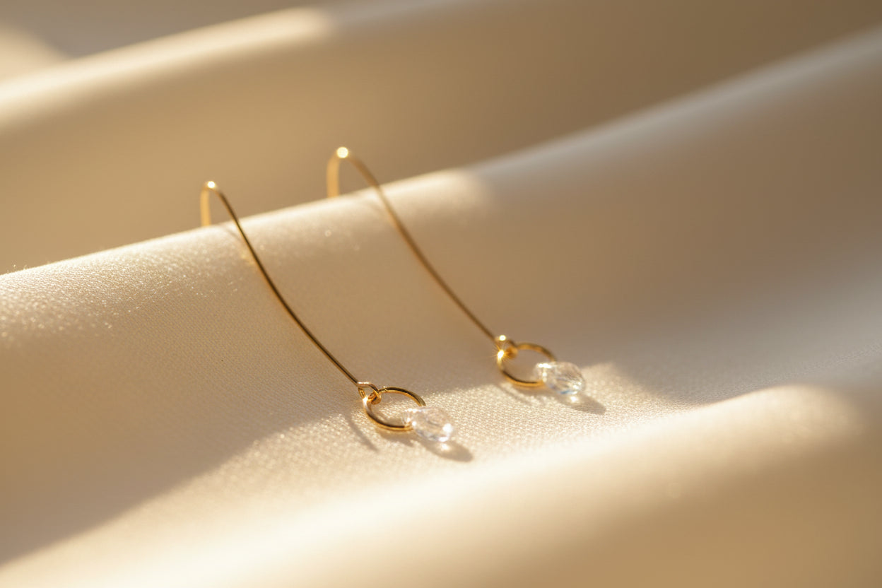 A pair of minimalist gold dangle earrings with thin vertical bars ending in small circular hoops, displayed on a clear acrylic stand against a light gray background.
