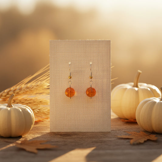 A pair of handmade dangle earrings with faceted orange beads and small gold beads on silver hooks, displayed on a clear plastic stand against a gray background.