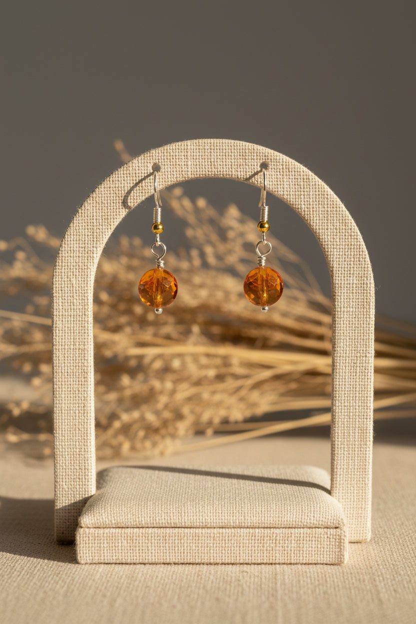 A pair of handmade dangle earrings with faceted orange beads and silver hooks, displayed on a clear plastic stand against a neutral gray background.