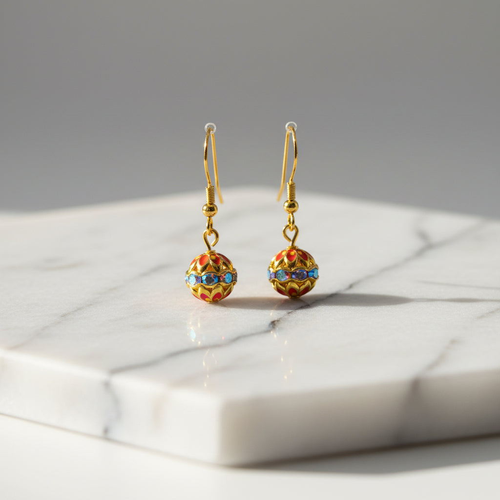 A pair of gold dangle earrings with small, colorful enamel-covered spherical charms, displayed on a clear acrylic stand against a neutral gray background.