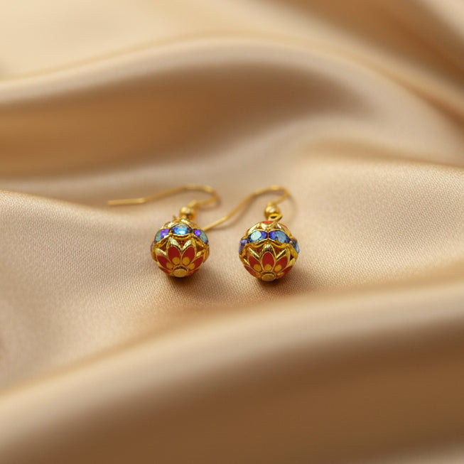 A pair of gold dangle earrings with colorful enamel ball charms, displayed on a clear acrylic stand against a neutral gray background.