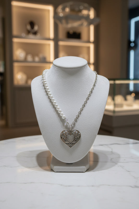 A pearl necklace with a silver heart-shaped pendant displayed on a white jewelry bust. The background shows a softly blurred boutique interior with warm lighting and shelving.