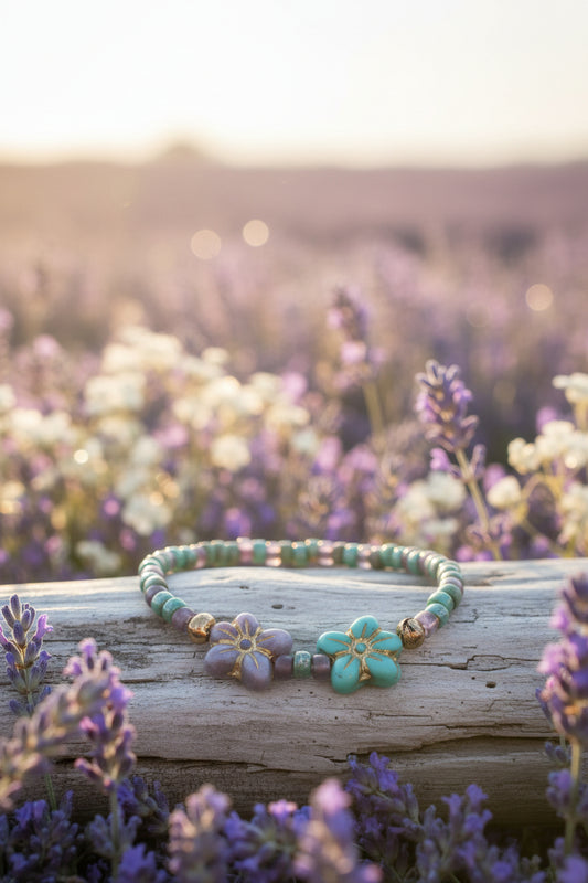 A beaded stretch bracelet featuring green and purple beads, two large flower-shaped beads (one purple, one turquoise with gold trim), and small gold spacer beads, displayed on a light gray surface.