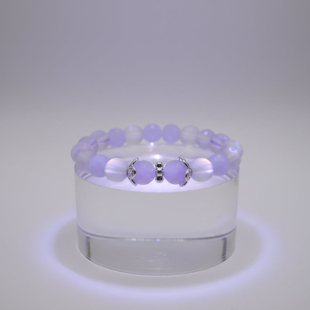 A beaded stretch bracelet featuring light purple and white beads, with two central silver-toned spacer beads flanking a round light purple bead, displayed on a neutral gray background.