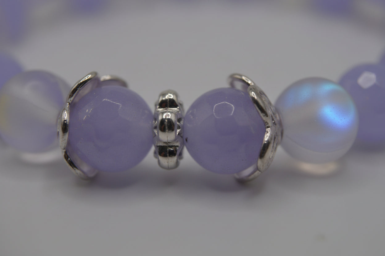 A beaded stretch bracelet featuring light purple and iridescent white beads, accented by two round purple beads set in silver-toned metal at the front, displayed on a neutral gray background.