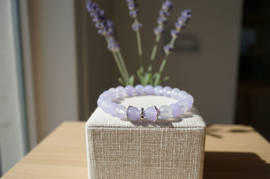 A beaded stretch bracelet featuring light purple and milky white round beads, accented by two larger lavender beads with silver-toned spacers in the front center, displayed on a neutral light gray background.