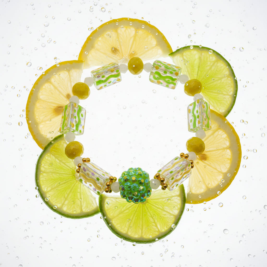 A handmade beaded stretch bracelet featuring translucent green and yellow rectangular beads, small yellow beads, and a central green glittery ball charm, displayed on a neutral gray background.