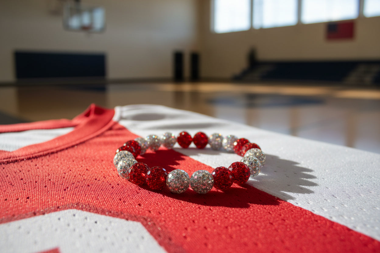 A beaded stretch bracelet with alternating red and silver glittery beads resting on a red and white basketball jersey in a blurred gymnasium setting.