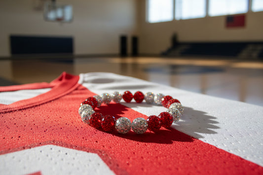 A beaded stretch bracelet with alternating red and silver glittery beads resting on a red and white basketball jersey in a blurred gymnasium setting.
