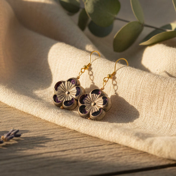 A pair of flower-shaped drop earrings with gold fishhook wires and dark purple petals, displayed on a clear acrylic stand against a neutral gray background.