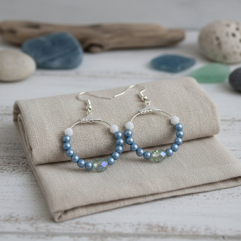 A pair of handmade hoop earrings with blue beads, white spacer beads, and a small clear faceted bead, hanging from silver-toned fishhook wires against a neutral gray background.