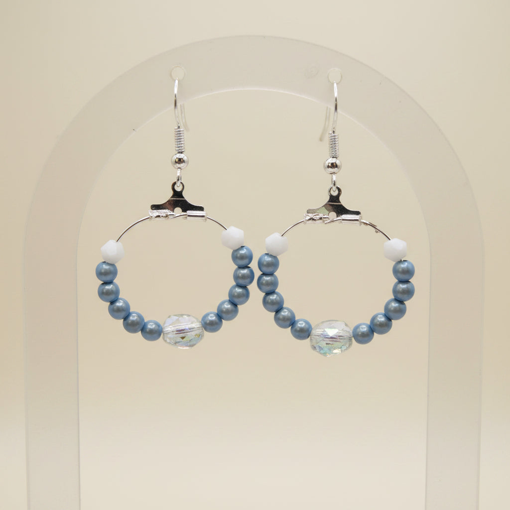 A pair of handmade hoop earrings with light blue beads, white spacer beads, and a small iridescent crystal accent, hanging from silver fishhook wires against a neutral gray background.
