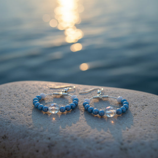 A pair of handmade hoop earrings with light blue beads, white spacer beads, and silver-toned metal hooks, displayed on a clear stand against a neutral gray background.