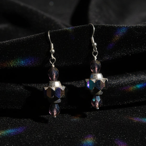 A pair of dangling earrings with silver-toned hooks and faceted beads in purple, black, and iridescent AB color, displayed on a clear stand against a light gray background.
