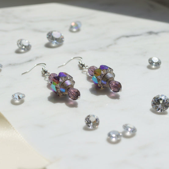 A pair of handmade dangle earrings with faceted purple beads and iridescent multicolor crystal clusters, hanging from silver fishhook wires on a clear display stand against a light gray background.