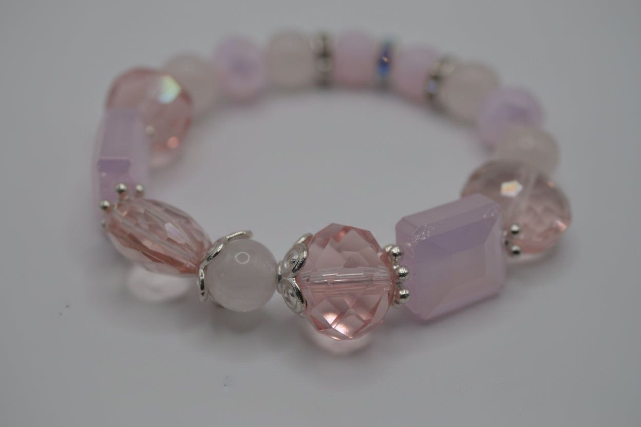 A beaded stretch bracelet featuring light pink and white beads, including faceted crystal beads, a rectangular pink bead, and small silver spacer rings, displayed on a neutral gray background.