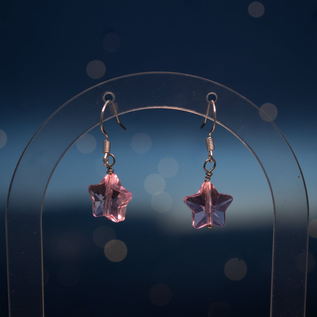 A pair of handmade dangle earrings featuring light pink star-shaped glass pieces suspended from silver fishhook wires, displayed on a clear acrylic stand against a neutral gray background.