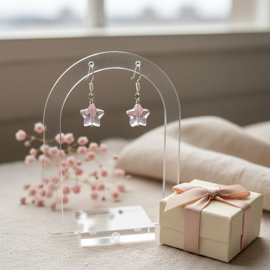 A pair of handmade dangle earrings featuring irregular, translucent light pink glass chips suspended from silver fishhook wires, displayed on a clear acrylic stand against a neutral gray background.