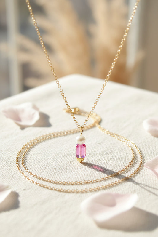 A delicate gold chain necklace with a pendant featuring a white bead and a pink faceted crystal bead, displayed on a black velvet jewelry bust.