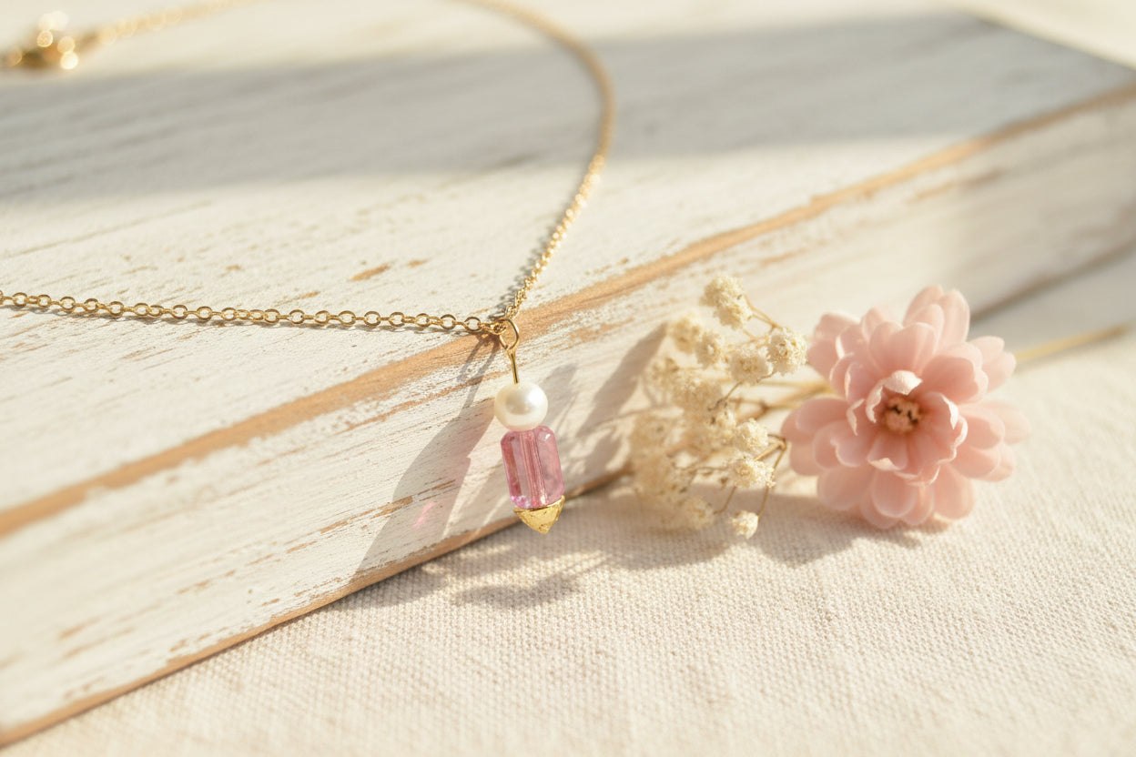 A delicate gold chain necklace displayed on a black velvet jewelry bust. The necklace features a central pendant with a small white bead and a faceted pink crystal hanging from gold-toned settings.