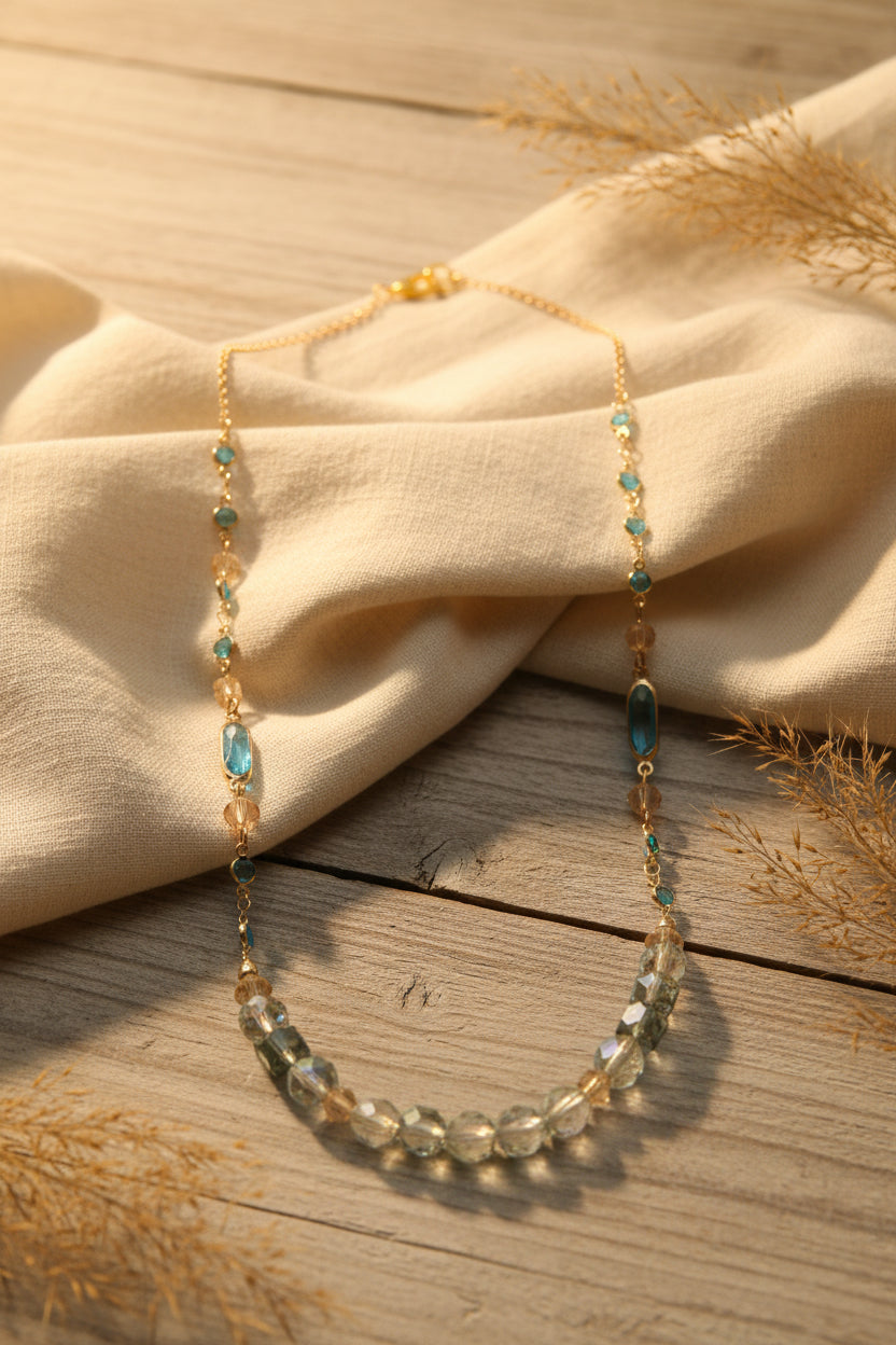 A delicate gold-tone beaded necklace featuring faceted iridescent beads, small turquoise blue beads, and blush pink beads, displayed against a neutral gray background.
