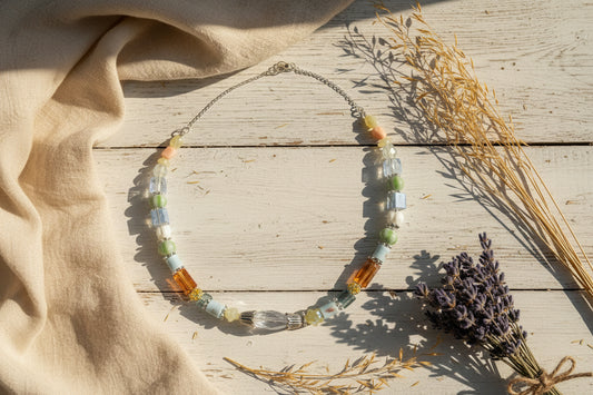 A beaded necklace featuring a mix of colorful glass beads in blue, green, orange, yellow, and white, accented by silver-toned metal spacers and a large clear faceted bead at the center, displayed on a neutral light gray background.
