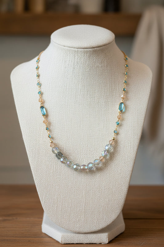 A gold-tone beaded necklace featuring faceted clear beads, iridescent green beads, and oval turquoise-blue beads, displayed against a plain light gray background.