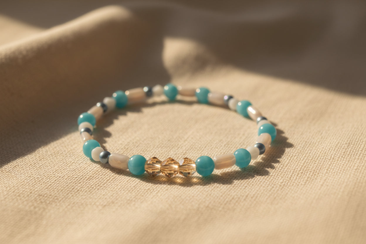 A beaded stretch bracelet featuring turquoise blue beads, white cylindrical beads, small metallic silver beads, and a central gold-toned geometric charm, displayed on a neutral gray background.