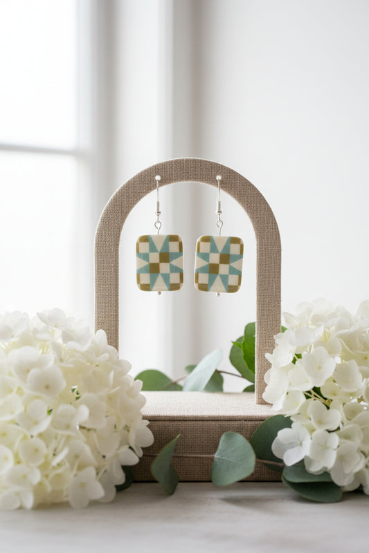 A pair of rectangular drop earrings with a geometric starburst pattern in sage green, light blue, and cream, suspended from silver fishhook wires on a clear display stand.