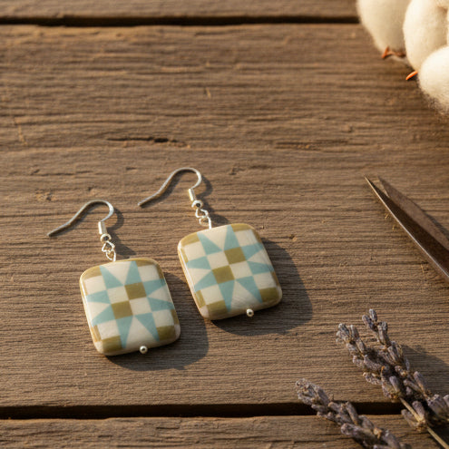 A pair of rectangular drop earrings with a geometric starburst pattern in sage green, light blue, and cream, suspended from silver fishhook wires against a neutral gray background.