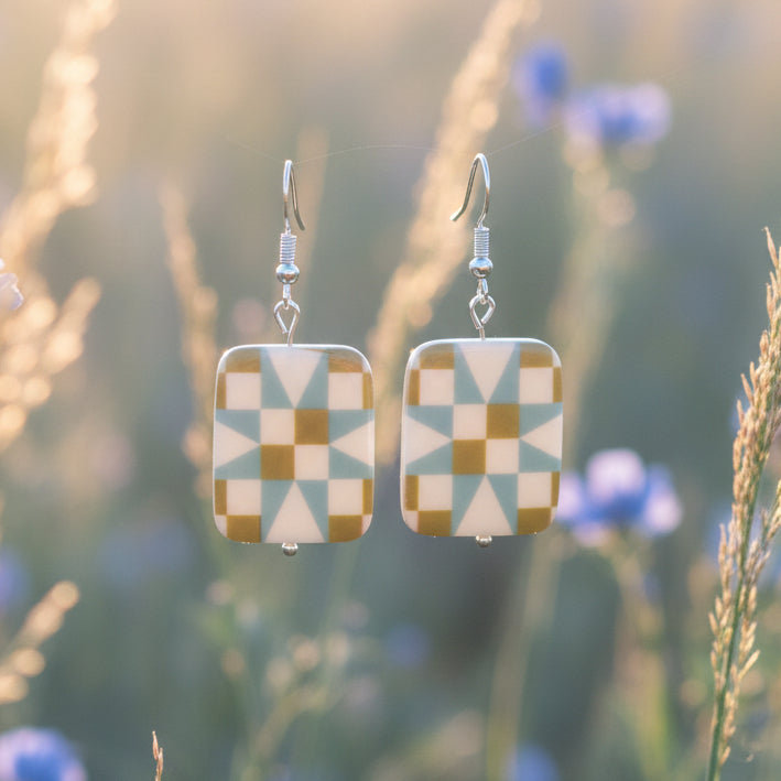 A pair of rectangular drop earrings with a geometric starburst pattern in sage green, light blue, and mustard yellow on a white background, hanging from silver fishhook wires.