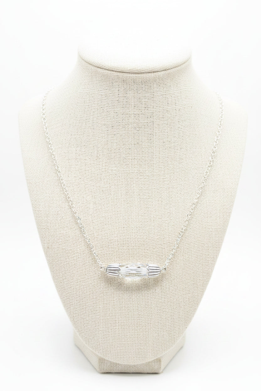 A silver chain necklace with a small, horizontally oriented oval pendant featuring a clear, iridescent center, displayed on a black velvet jewelry bust against a white background.