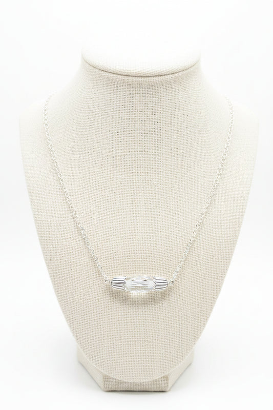 A silver chain necklace with a small, horizontally oriented oval pendant featuring a clear, iridescent center, displayed on a black velvet jewelry bust against a white background.