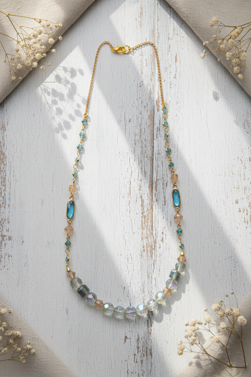 A delicate gold-tone beaded necklace featuring light blue oval beads, faceted clear beads, and small iridescent green accents, displayed against a neutral gray background.