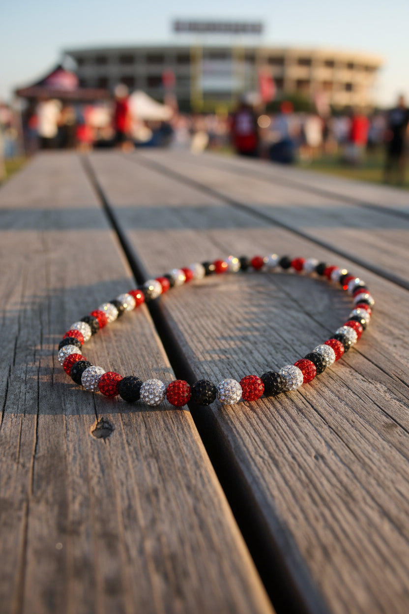 A beaded necklace with alternating red, black, and silver rhinestone-covered round beads, arranged in an oval shape on a plain light gray background.