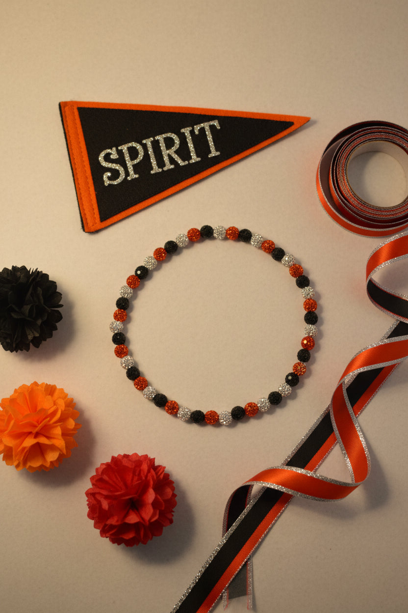 A flat-lay arrangement of Halloween-themed decorations on a light beige background, including a black and orange pennant with the word 'SPIRIT', a beaded bracelet, orange and black pom-poms, and orange, black, and silver ribbon.
