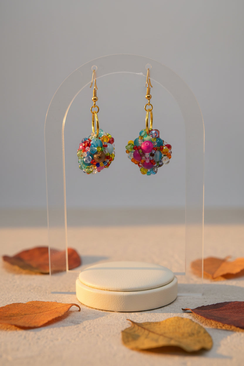 A pair of handmade beaded dangle earrings with gold fishhook wires and colorful glass beads, displayed on a clear acrylic stand against a neutral gray background.