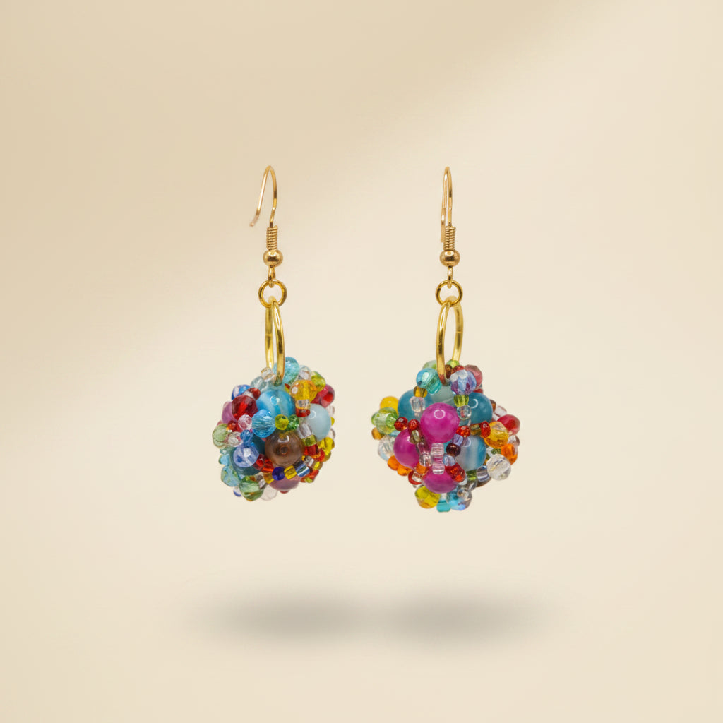A pair of handmade beaded dangle earrings with gold fishhook wires and gold O-rings, featuring colorful glass beads in pink, blue, yellow, and green, displayed on a clear acrylic stand against a neutral gray background.