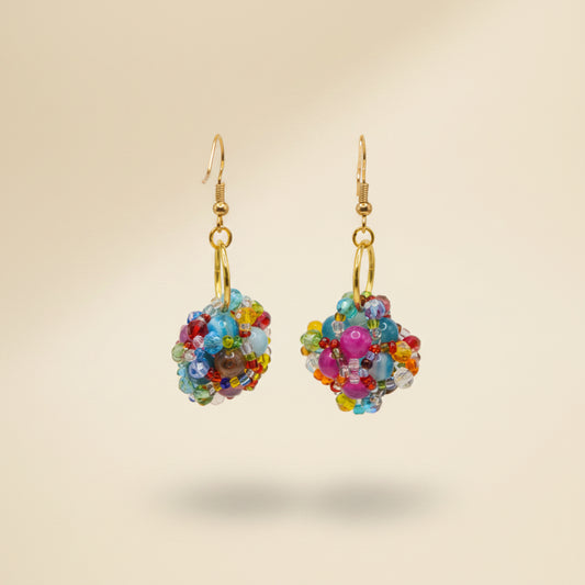 A pair of handmade beaded dangle earrings with gold fishhook wires and gold O-rings, featuring colorful glass beads in pink, blue, yellow, and green, displayed on a clear acrylic stand against a neutral gray background.