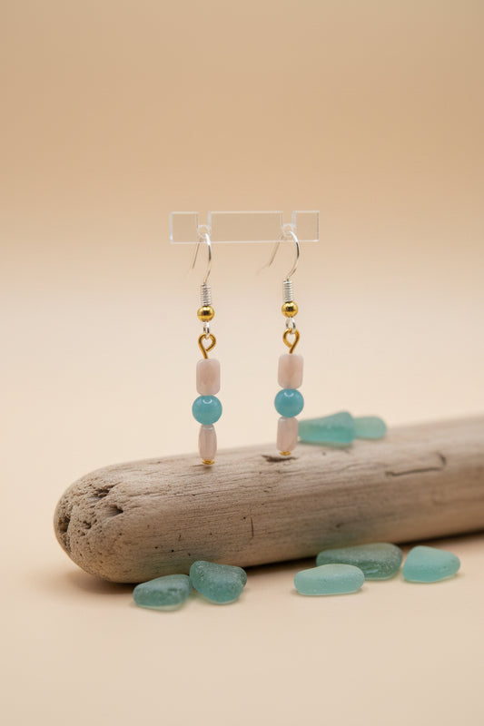 A pair of handmade dangle earrings displayed on a clear acrylic stand against a neutral gray background. The earrings feature silver-toned fishhook wires, small gold spacer beads, a light pink cylindrical bead, a round light blue bead, and a smaller pink cylindrical bead at the bottom.