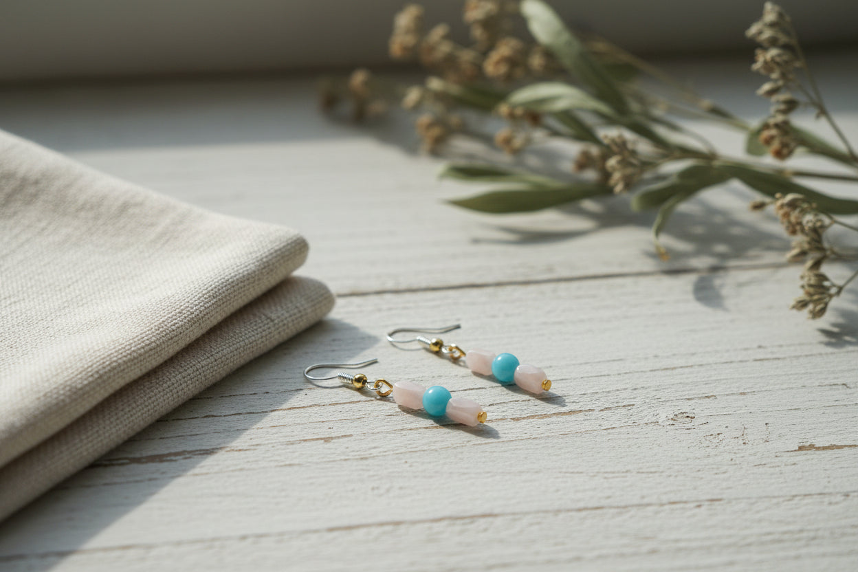 A pair of handmade beaded dangle earrings hanging from a clear acrylic display stand against a neutral gray background. The earrings feature silver-toned fishhook wires, small gold-toned beads, a pale pink rectangular bead, a round light blue bead, and a small gold-toned bead at the bottom.
