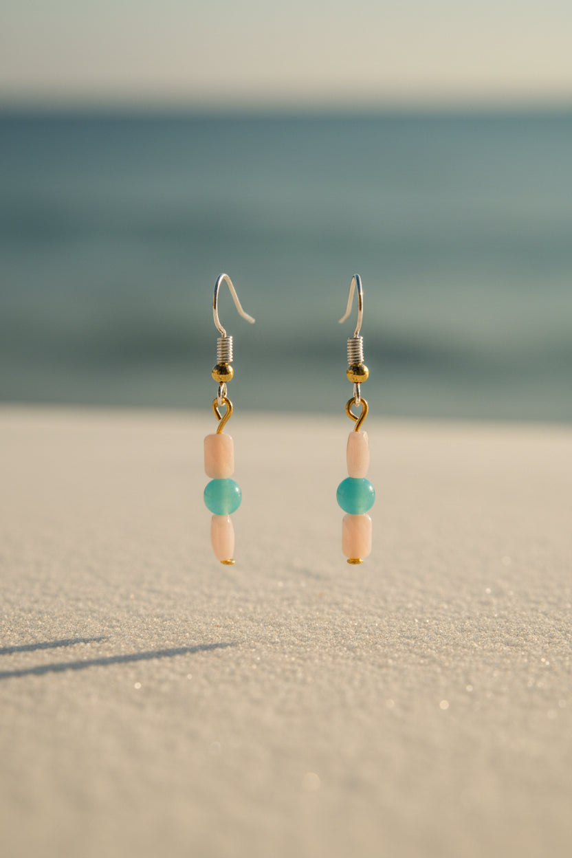A pair of handmade dangle earrings with light pink beads, a turquoise blue bead, and gold-toned metal hooks, displayed on a clear acrylic stand against a light gray background.