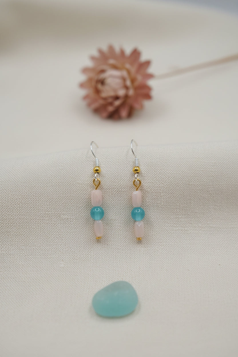 A pair of handmade beaded dangle earrings displayed on a clear acrylic stand against a neutral gray background. The earrings feature gold-tone metal hooks and connectors, pink glass beads, a light blue bead, and a small gold spacer bead.