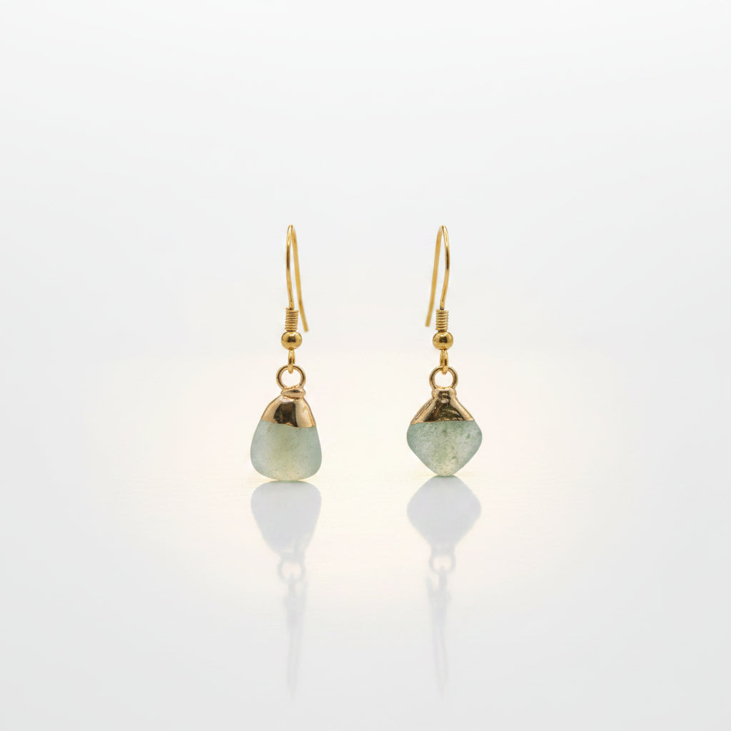 A pair of gold dangle earrings with irregular light green stone pendants, displayed on a clear acrylic stand against a neutral gray background.