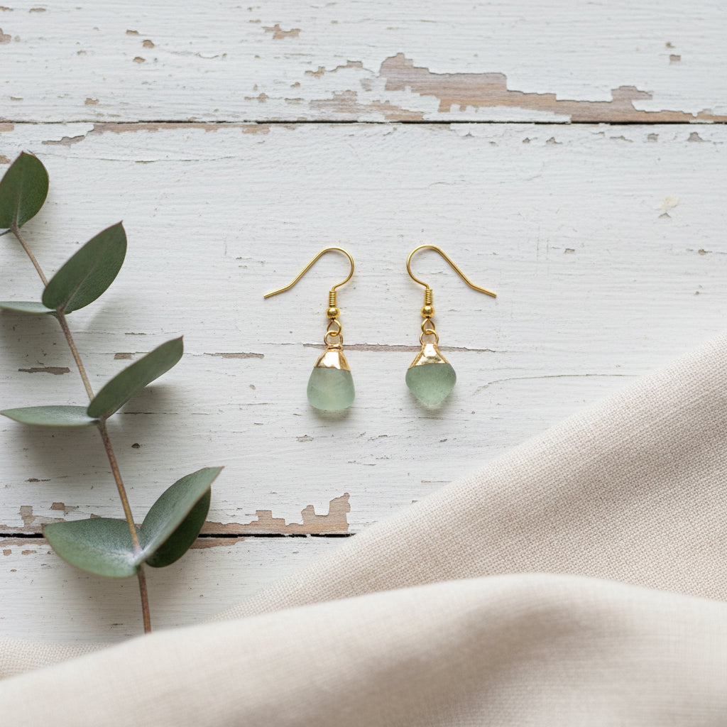 A pair of gold drop earrings with irregular light green gemstone pendants, displayed on a clear acrylic stand against a neutral gray background.