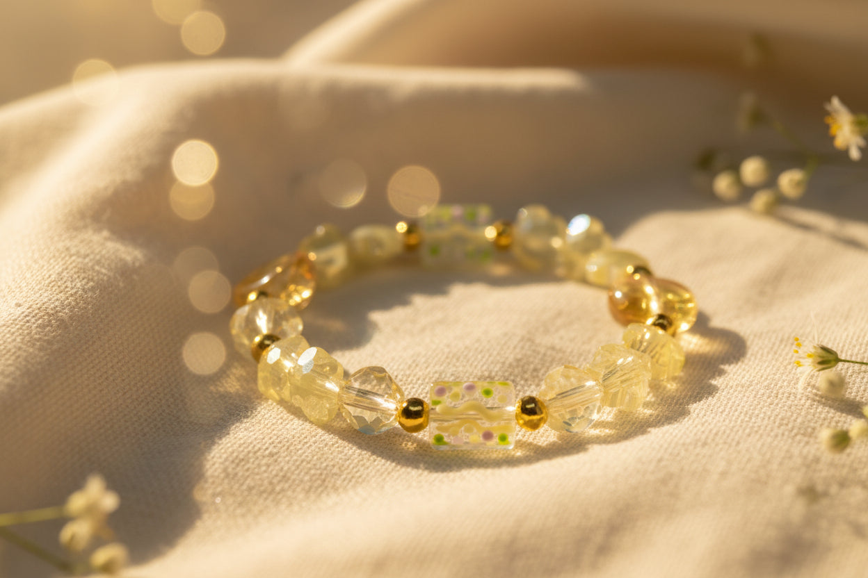 A beaded stretch bracelet featuring pale yellow translucent beads, textured flower-shaped beads, round amber-yellow beads, and a central rectangular glass bead with wavy yellow and green patterns, accented by small gold spacer beads, shown on a plain light gray background.