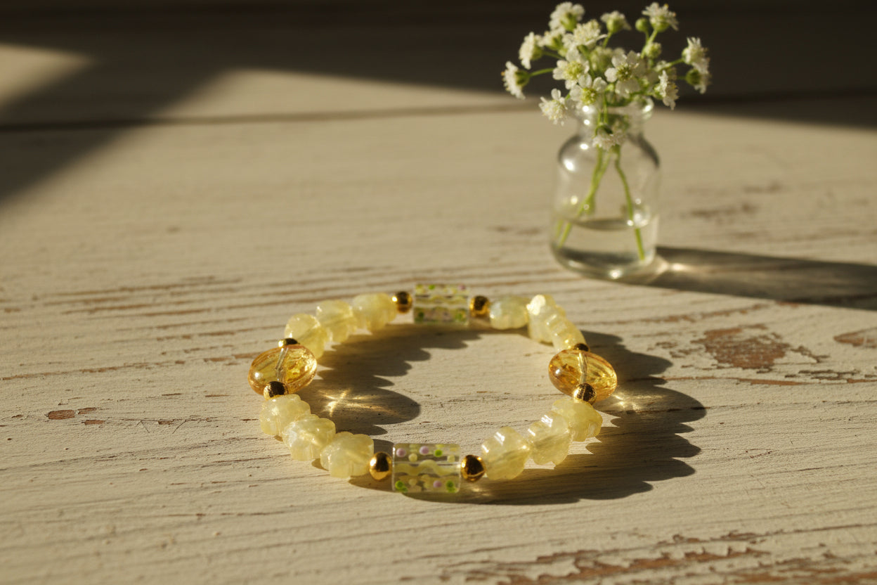 A beaded stretch bracelet featuring pale yellow translucent beads, textured flower-shaped beads, round amber-toned beads, and a central rectangular bead with wavy yellow lines and small multicolor speckles, separated by small gold spacer beads, on a plain light gray background.