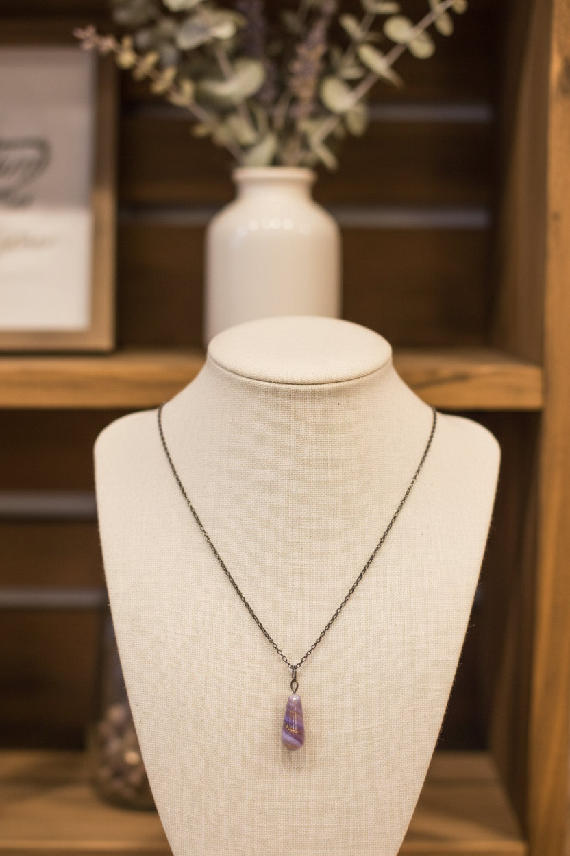 A silver chain necklace with a teardrop-shaped pendant featuring horizontal lavender, purple, and orange stripes, displayed on a black velvet jewelry bust.